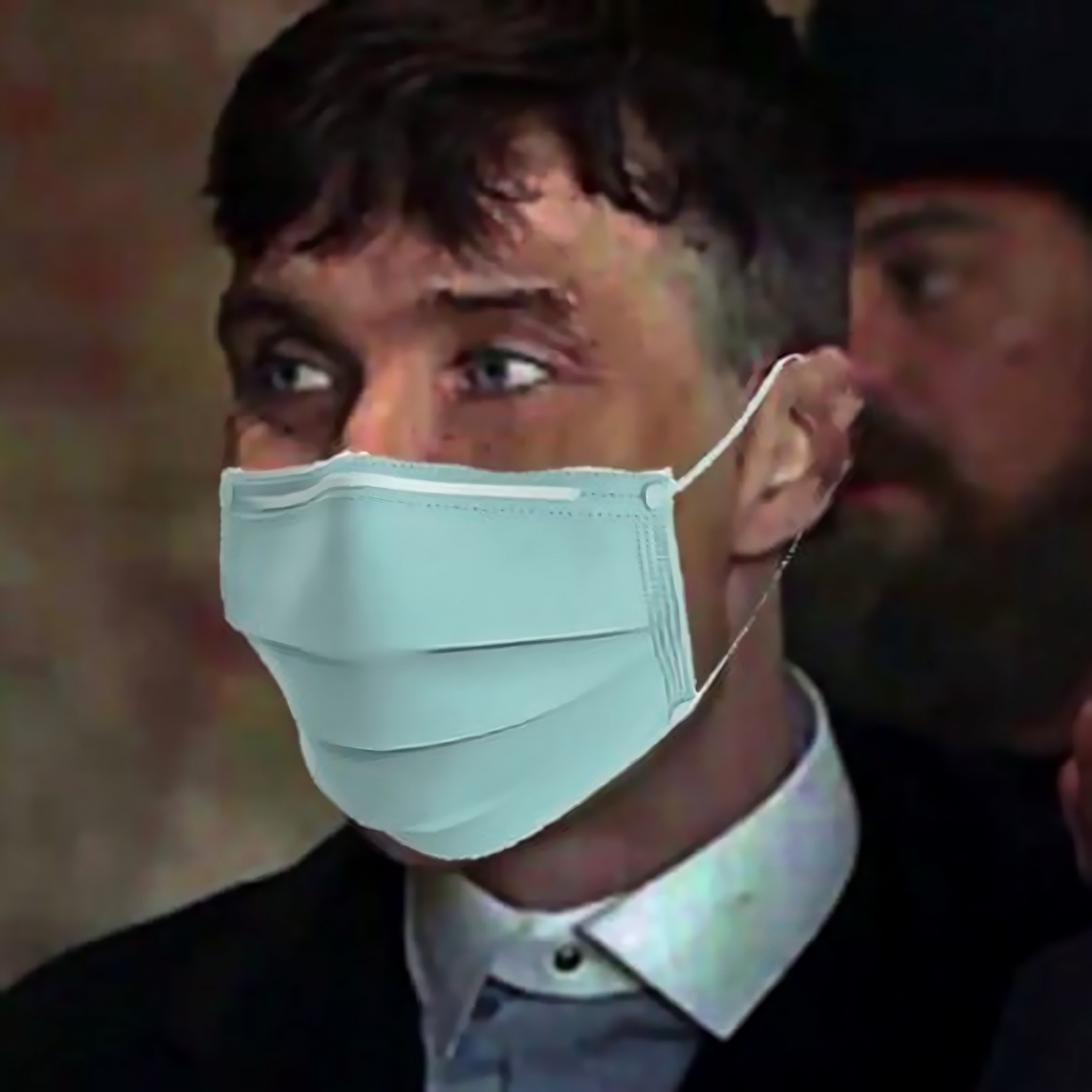 Adam Mourn in a mint surgical mask and dark suit, looking off frame, a bearded man behind him